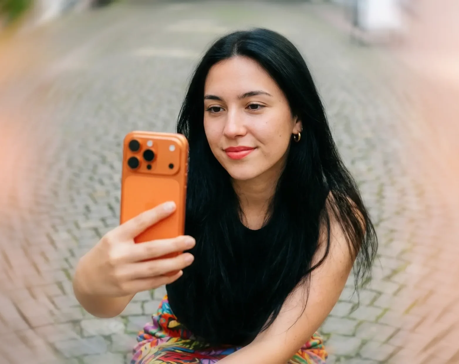 A frontal editorial portrait of a swiss gen z girl with long black hair, gold earrings and light red lipstick,, shot on a 85 mm lens under soft light. She wears a colorful skirt Holding an orange iphone 17 pro and taking a selfie f. Behind her a the streets of Aarau, evenly lit to make her facial features stand out clearly without glare. Her satisfied expression and skin texture are sharp and realistic. Lens flares and fine film grain give the photo an authentic analog feel.