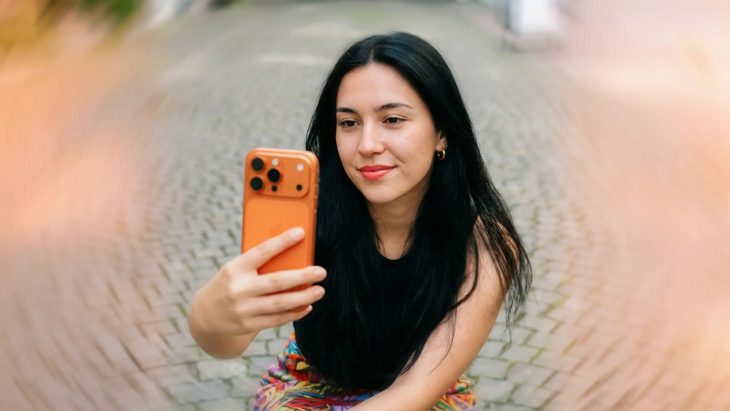 A frontal editorial portrait of a swiss gen z girl with long black hair, gold earrings and light red lipstick,, shot on a 85 mm lens under soft light. She wears a colorful skirt Holding an orange iphone 17 pro and taking a selfie f. Behind her a the streets of Aarau, evenly lit to make her facial features stand out clearly without glare. Her satisfied expression and skin texture are sharp and realistic. Lens flares and fine film grain give the photo an authentic analog feel.